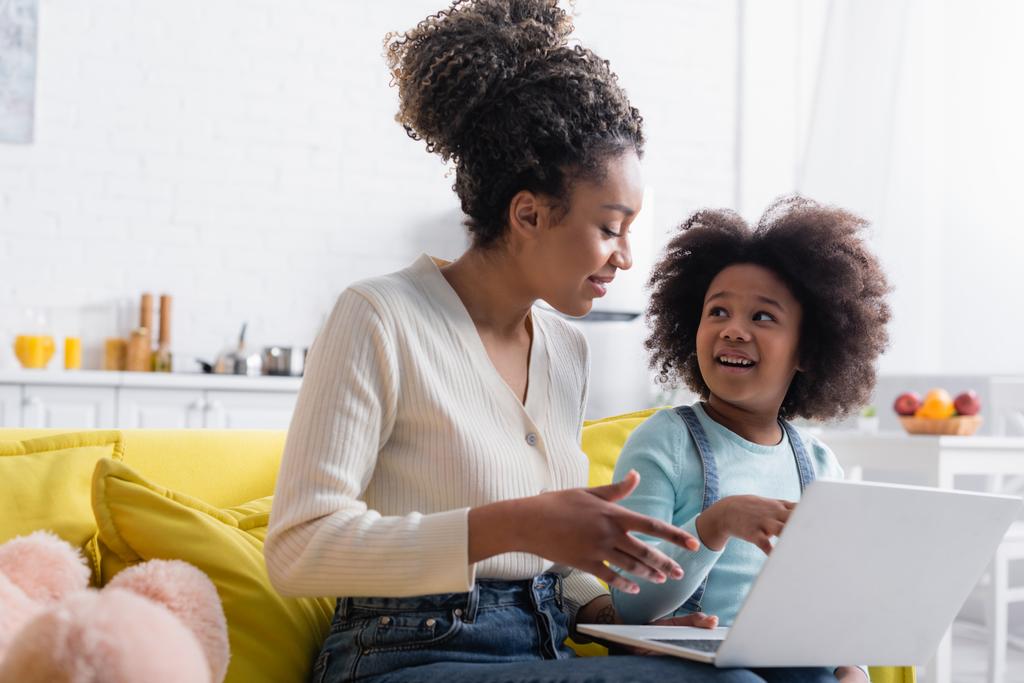 Gülümseyen Afro-Amerikan anne ve kızı dizüstü bilgisayarı işaret ederken birbirlerine bakıyorlar. - Fotoğraf, Görsel