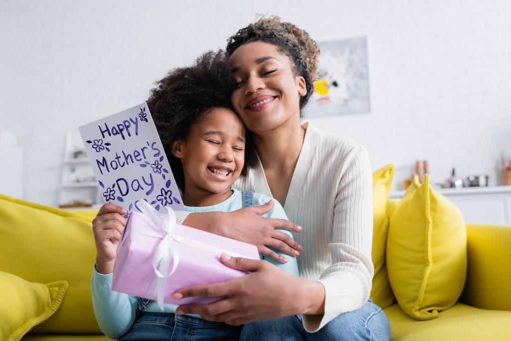 ευχαριστημένη Αφροαμερικανή γυναίκα που κρατάει κουτί δώρου και αγκαλιάζει την κόρη της με ευχάριστες κάρτες ημέρας μητέρων - Φωτογραφία, εικόνα