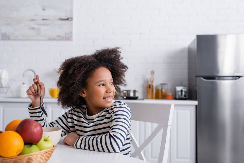 joyful african american girl holding spoon while looking away during breakfast in kitchen - Photo, Image