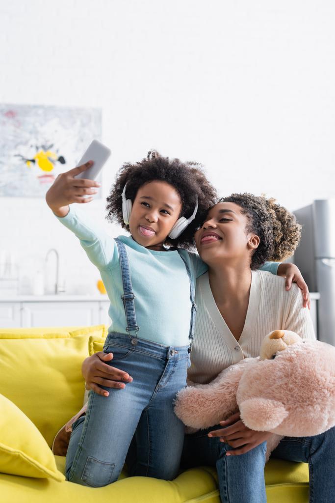 african american girl in headphones and woman with teddy bear taking selfie while sticking out tongues - Photo, Image