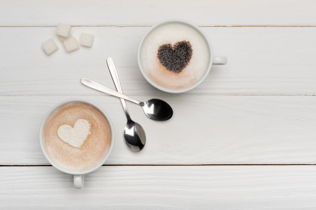 vrchní pohled na poháry s cappuccino a latté v blízkosti lžíce a kostky cukru na bílém dřevěném pozadí   - Fotografie, Obrázek
