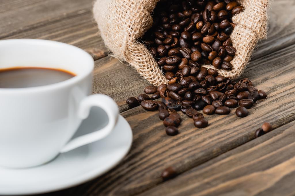 zsákzsák pörkölt kávébabbal a homályos csésze mellett, fa felületen  - Fotó, kép