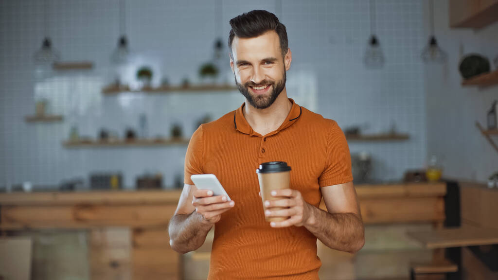 usmívající se muž pomocí smartphonu a držení kávy jít do kavárny - Fotografie, Obrázek