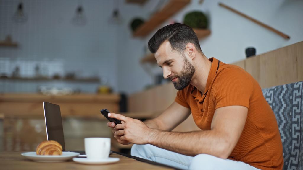 улыбающийся фрилансер на смартфоне рядом с ноутбуком и завтраком в кафе  - Фото, изображение