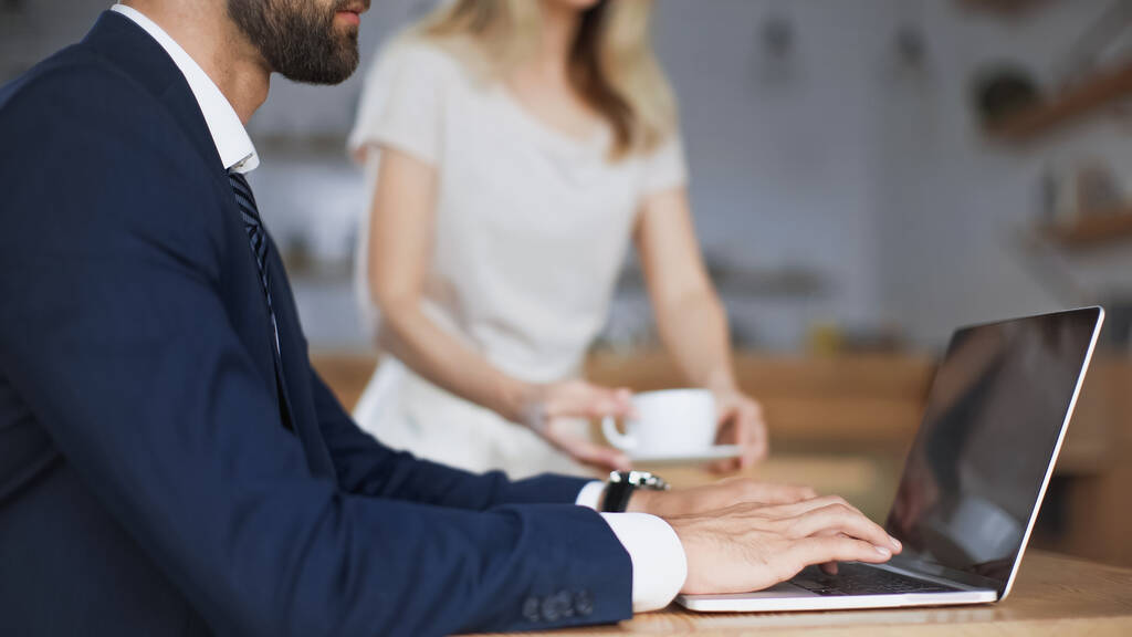 bijgesneden weergave van bebaarde zakenman typen op laptop in de buurt van vrouw met kopje op wazig achtergrond  - Foto, afbeelding