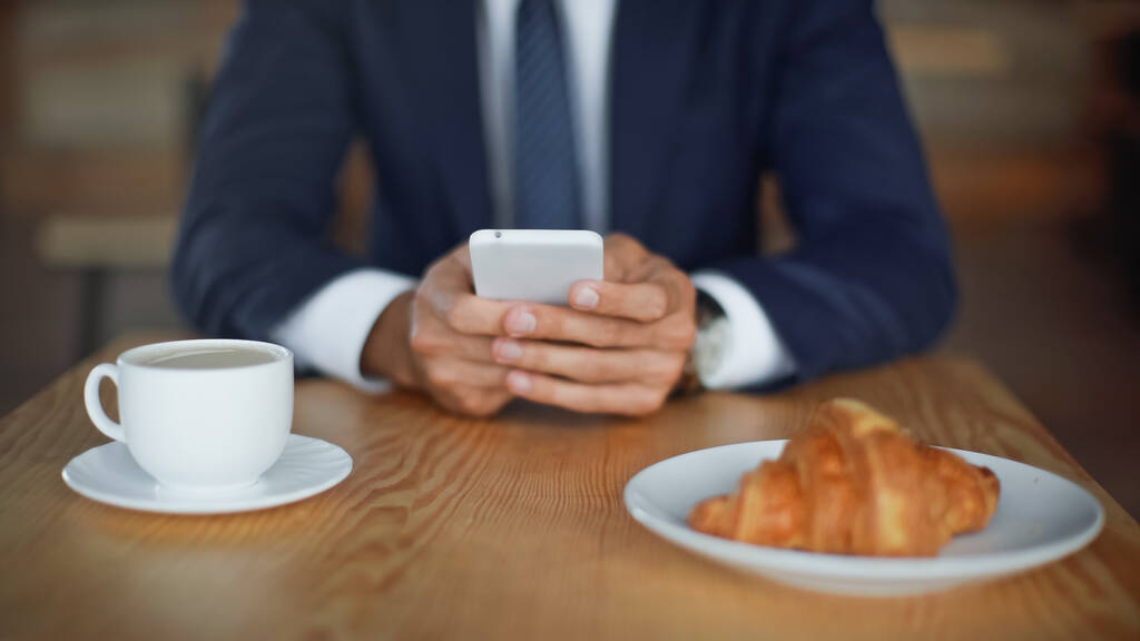 vista recortada de hombre de negocios utilizando teléfono inteligente cerca de croissant y café en la taza en primer plano borrosa  - Foto, Imagen