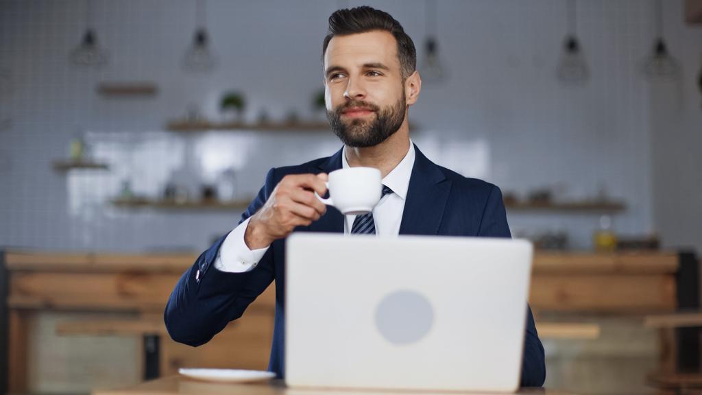 pleased man in suit holding cup of coffee near laptop in cafe - Photo, Image