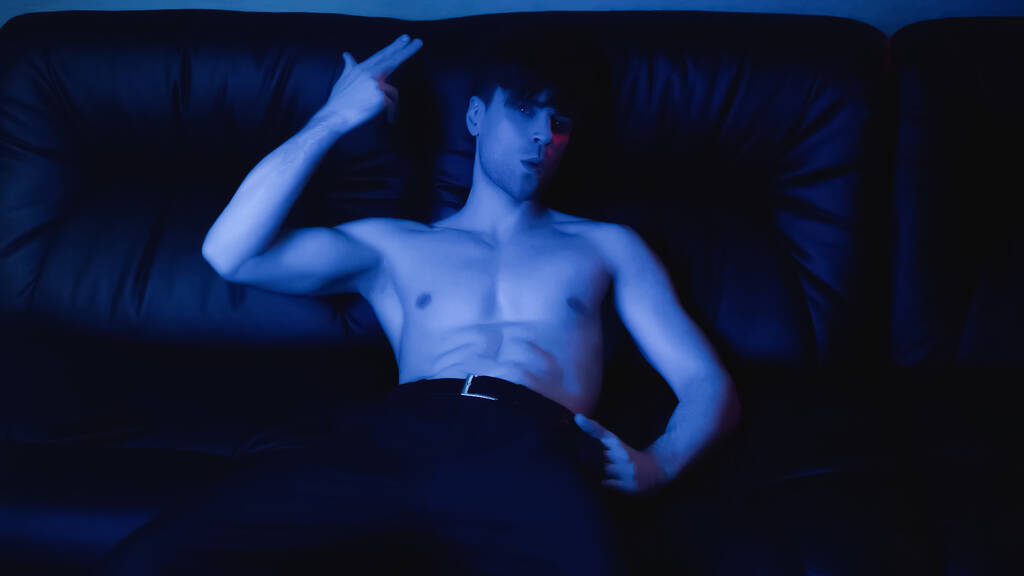 niebieskie oświetlenie na człowieka bez koszuli w spodniach pozowanie i gestykulacji podczas odpoczynku na czarnej kanapie  - Zdjęcie, obraz