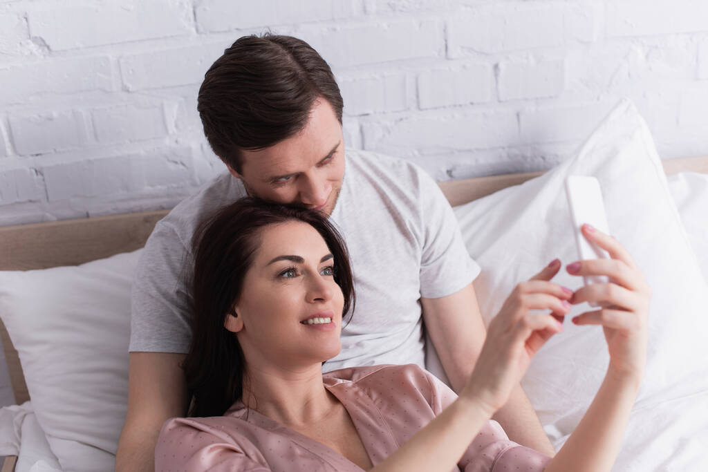 Usmívající se žena pomocí mobilu na rozmazané popředí v blízkosti manžela na posteli  - Fotografie, Obrázek