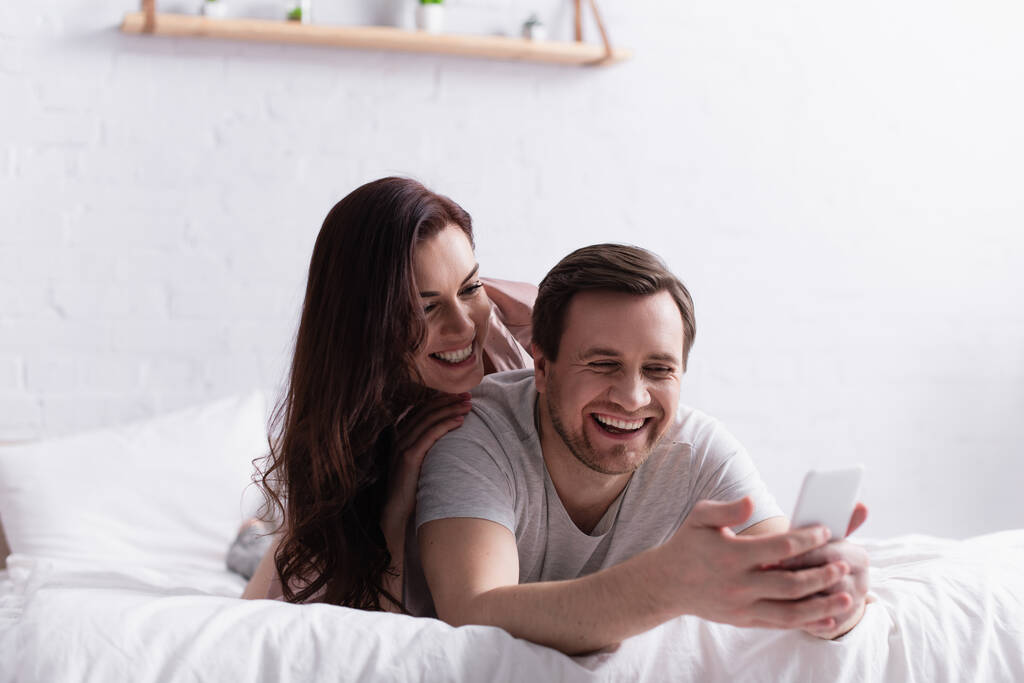 ベッドの上で明るい妻の近くにぼやけた前景にスマートフォンを使用して笑顔男  - 写真・画像