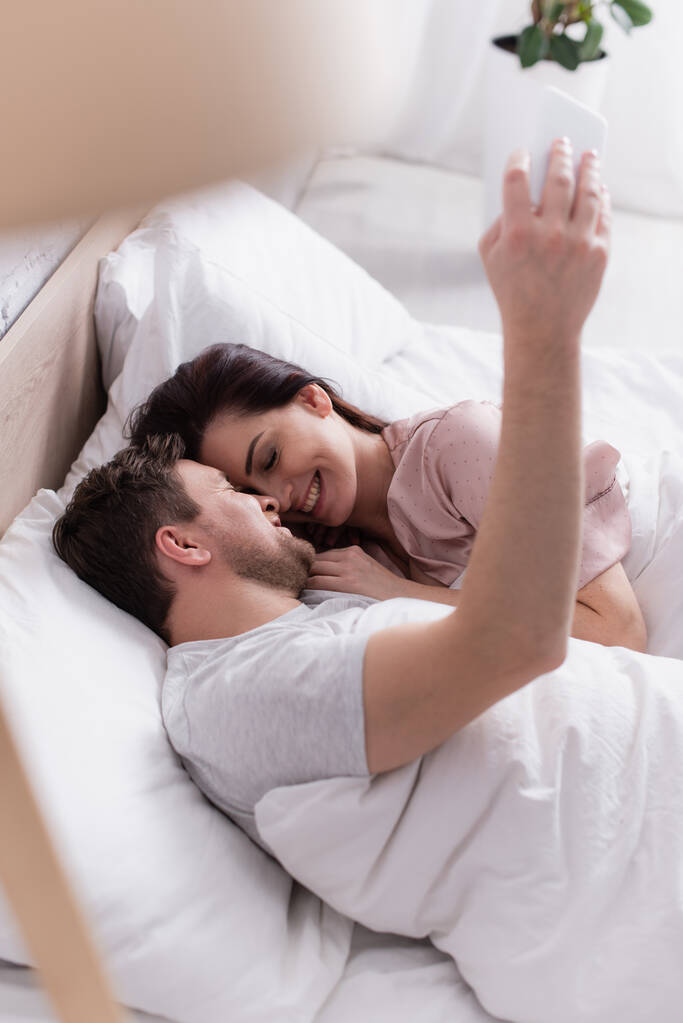 Sonriente pareja tomando selfie en el teléfono inteligente en la cama  - Foto, imagen