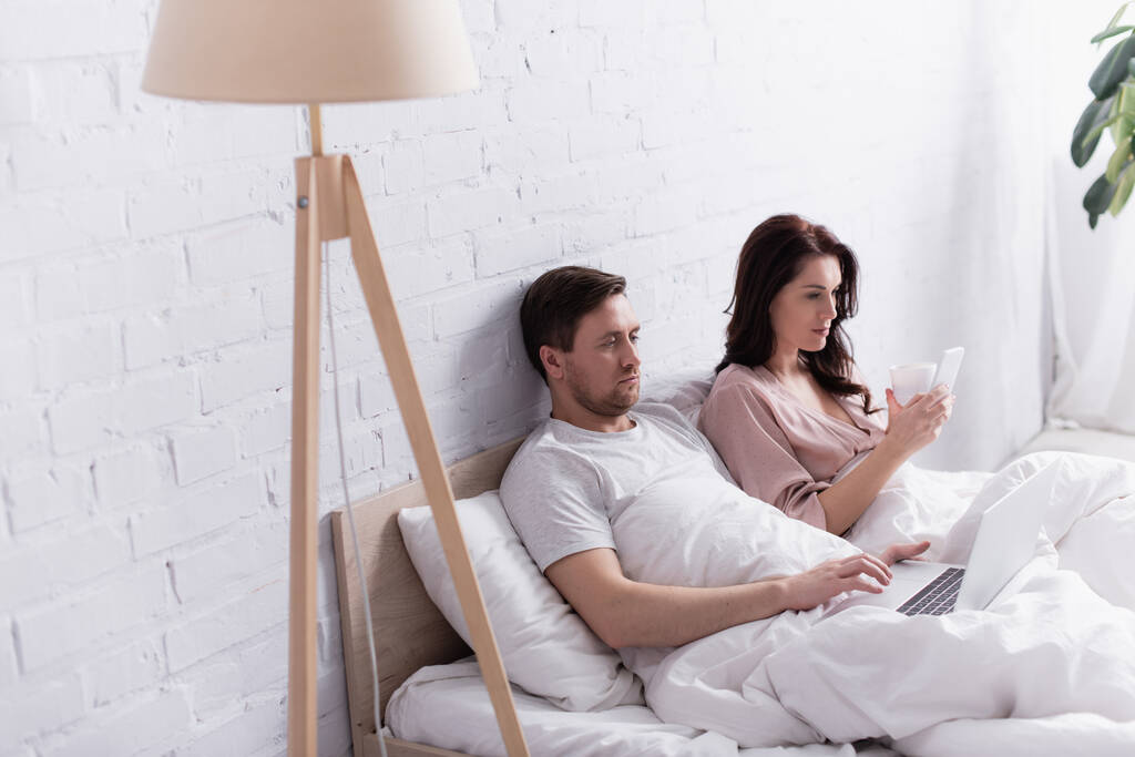 Мужчина с ноутбуком рядом с женой с чашкой и смартфоном в постели  - Фото, изображение