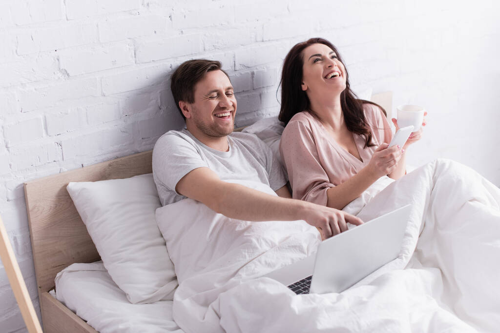 Cheerful couple with smartphone and laptop in bed  - Photo, Image