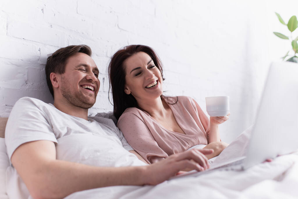 Χαμογελώντας άνθρωπος χρησιμοποιώντας φορητό υπολογιστή σε θολή πρώτο πλάνο κοντά στη σύζυγο με κύπελλο στο κρεβάτι  - Φωτογραφία, εικόνα