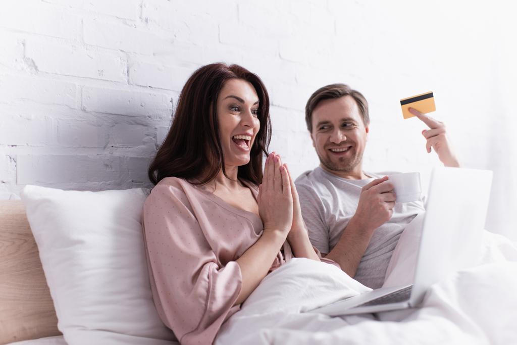 Positive Frau mit betenden Händen blickt auf Laptop neben Ehemann mit Kreditkarte und Tasse im Schlafzimmer  - Foto, Bild