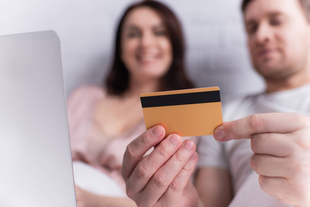 Кредитная карта в руках мужчины возле ноутбука и жены на размытом фоне  - Фото, изображение
