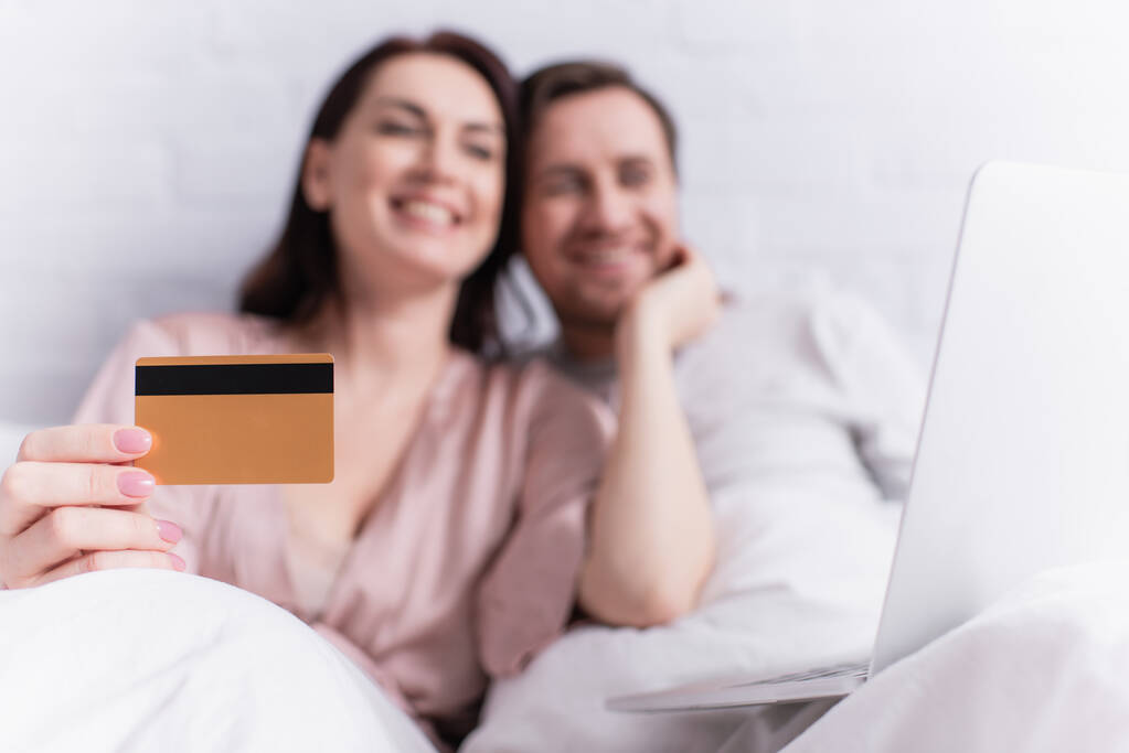 Carte de crédit en main de la femme floue près du mari et ordinateur portable sur le lit  - Photo, image