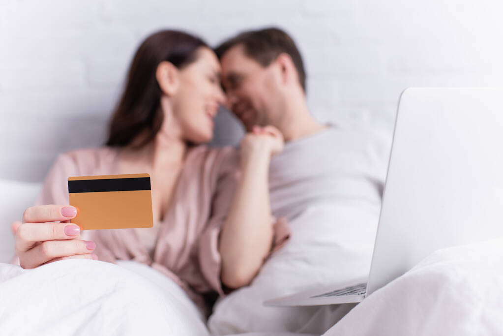 Laptop near woman holding credit card and looking at husband on blurred background on bed  - Photo, Image