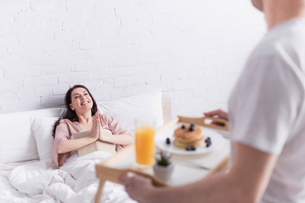 Усміхнена жінка з книгою, що показує, будь ласка, жест біля розмитого чоловіка зі сніданком у спальні
  - Фото, зображення