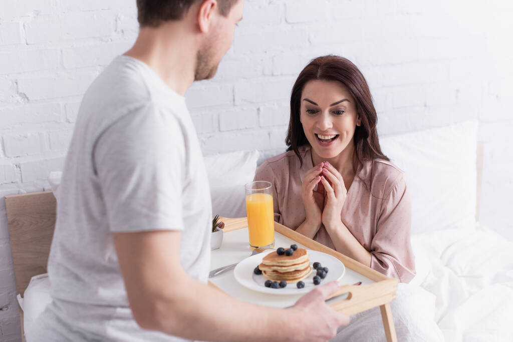 Szczęśliwa kobieta patrząc na smaczne naleśniki i sok pomarańczowy na tacy w pobliżu męża w sypialni  - Zdjęcie, obraz