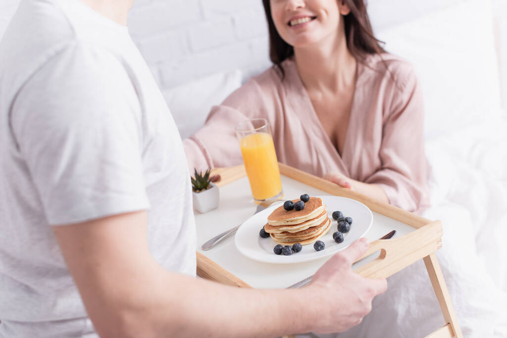 Cropped view of delicious pancakes and orange juice on tray near couple on blurred background in bedroom  - Photo, Image