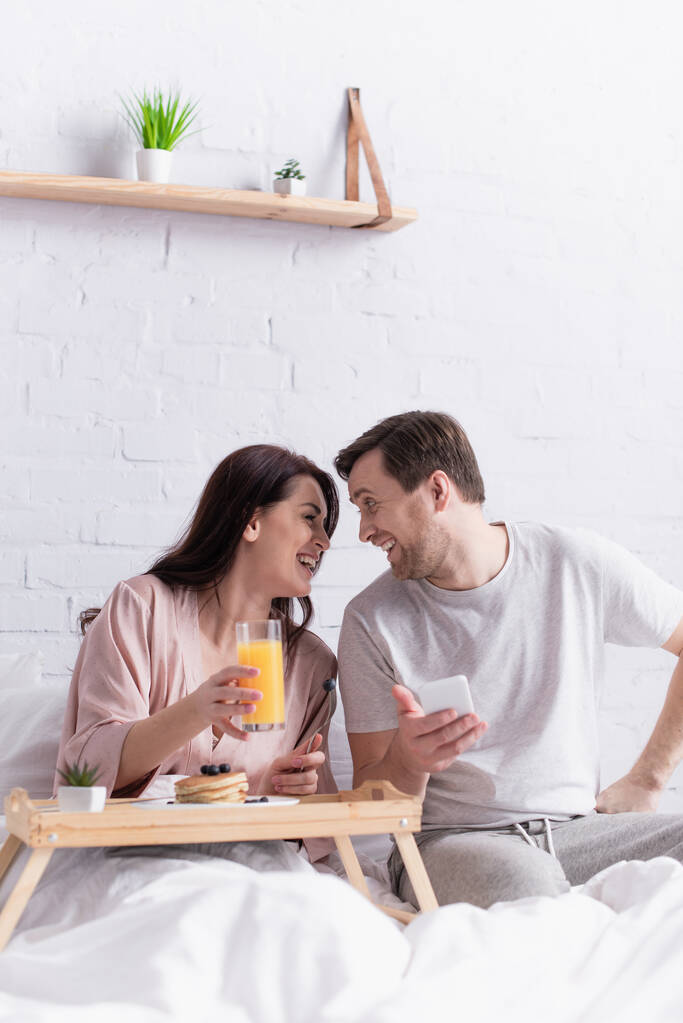 Mujer sonriente sosteniendo jugo de naranja cerca del desayuno y marido con teléfono inteligente  - Foto, imagen