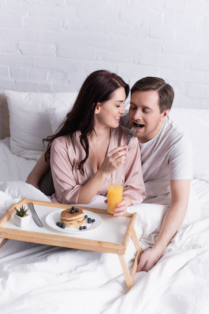 Femme souriante nourrissant son mari près des crêpes et du jus d'orange au lit  - Photo, image