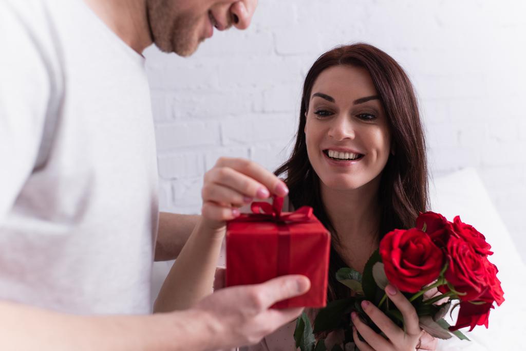 Χαρούμενη γυναίκα με τριαντάφυλλα που κρατά τόξο του παρόντος κοντά στο σύζυγό της σε θολή πρώτο πλάνο  - Φωτογραφία, εικόνα