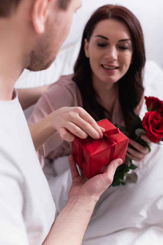 Mann hält Geschenk in der Nähe seiner Frau mit Rosen im Bett  - Foto, Bild