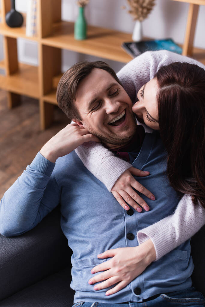 Brunette woman embracing cheerful husband on sofa at home  - Photo, Image