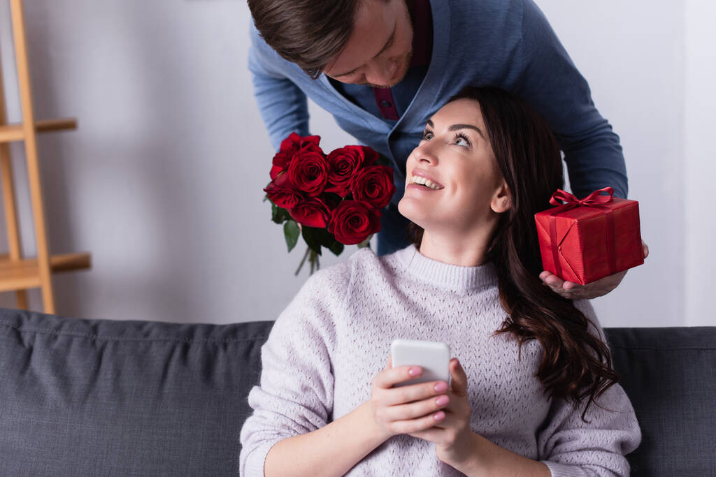 Usmívající se žena drží smartphone poblíž manžela s květinami a dárek  - Fotografie, Obrázek