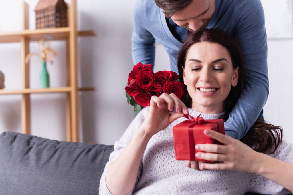 glimlachende vrouw houden aanwezig met strik in de buurt van echtgenoot met rode rozen  - Foto, afbeelding