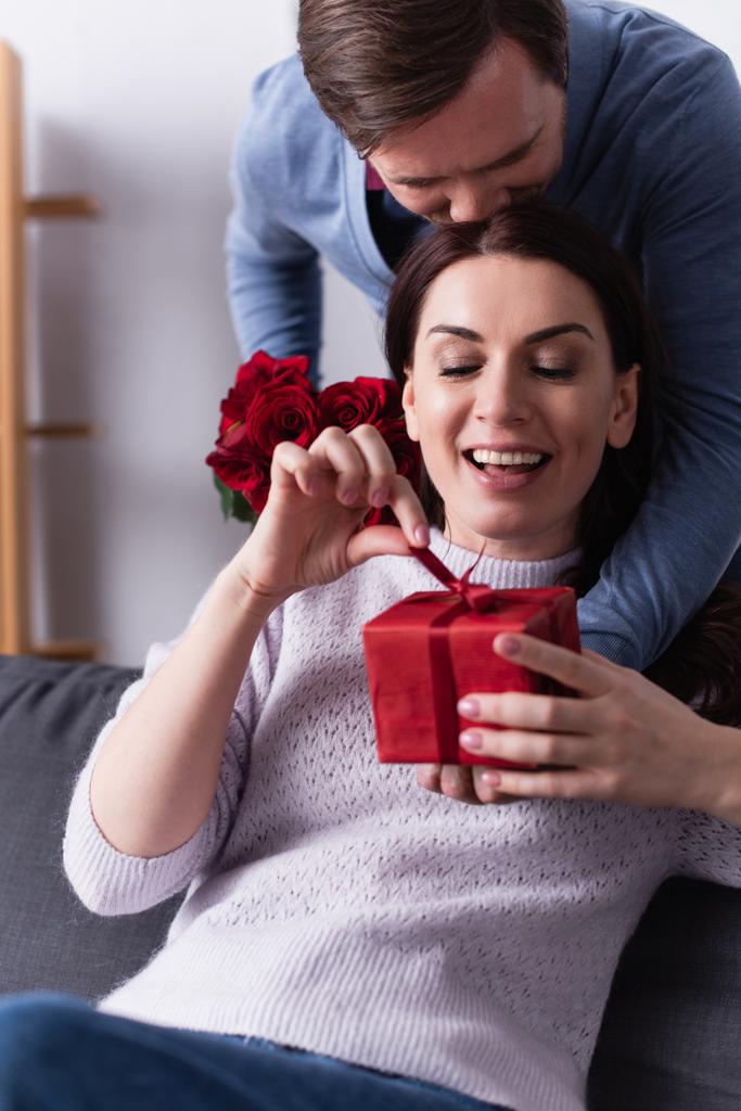 Mosolygó nő kezében íj ajándék doboz közelében férj rózsákkal otthon  - Fotó, kép