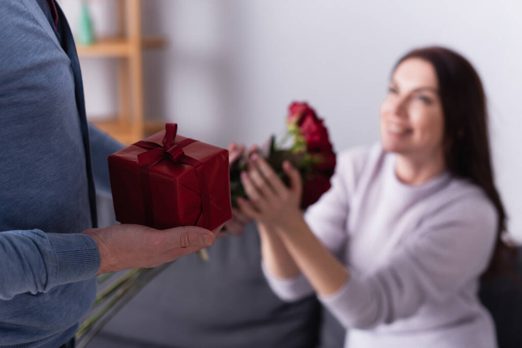 Άντρας κρατώντας κουτί δώρου κοντά στη σύζυγο με λουλούδια σε θολή φόντο  - Φωτογραφία, εικόνα