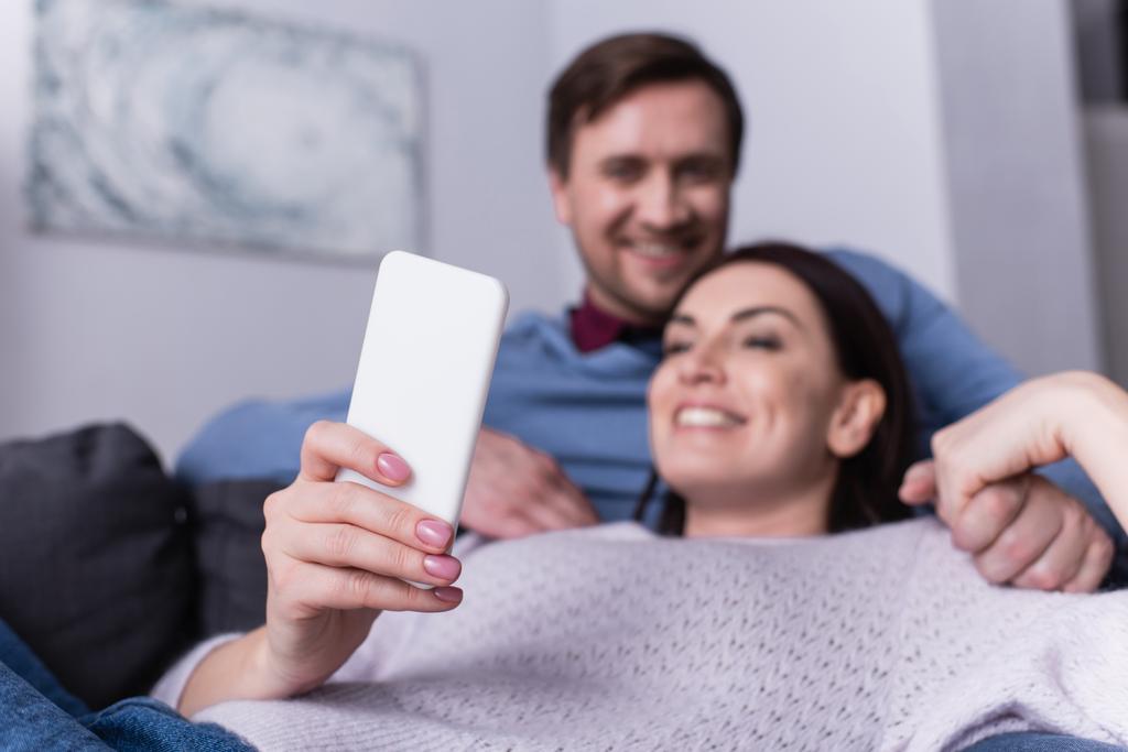 Smartphone en la mano de la mujer sonriente acostada en el sofá cerca del marido  - Foto, Imagen