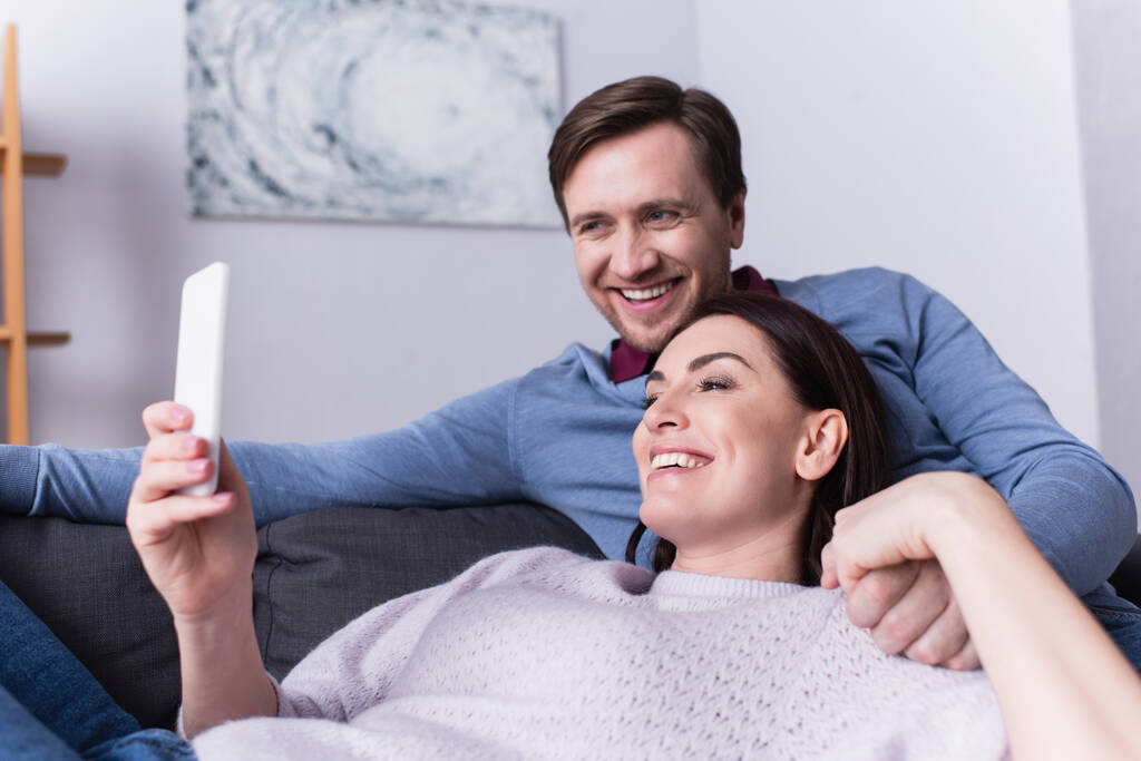 Hymyilevä aikuinen pari käyttää älypuhelinta sohvalla  - Valokuva, kuva