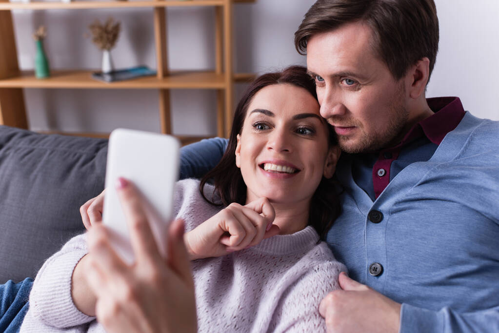 Mulher positiva segurando celular em primeiro plano borrado perto do marido na sala de estar  - Foto, Imagem