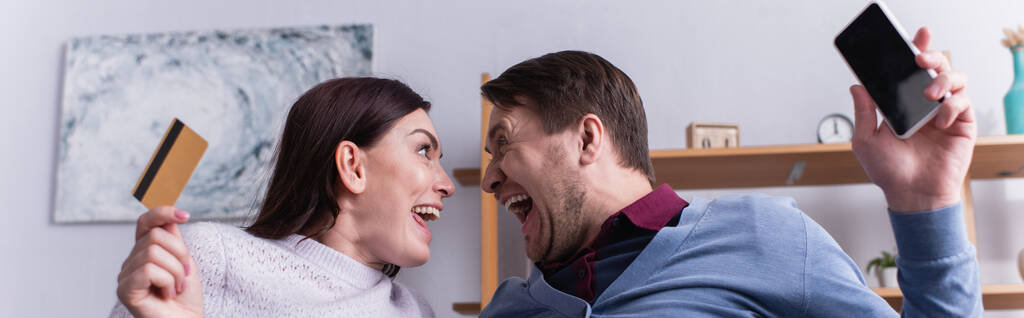 Úžasný pár s kreditní kartou a smartphone při pohledu na sebe doma, banner  - Fotografie, Obrázek