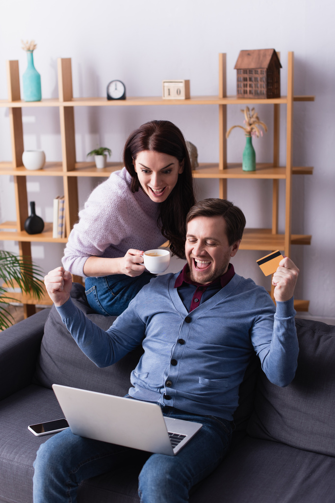 Homme excité tenant carte de crédit près d'un ordinateur portable et femme avec tasse à la maison  - Photo, image