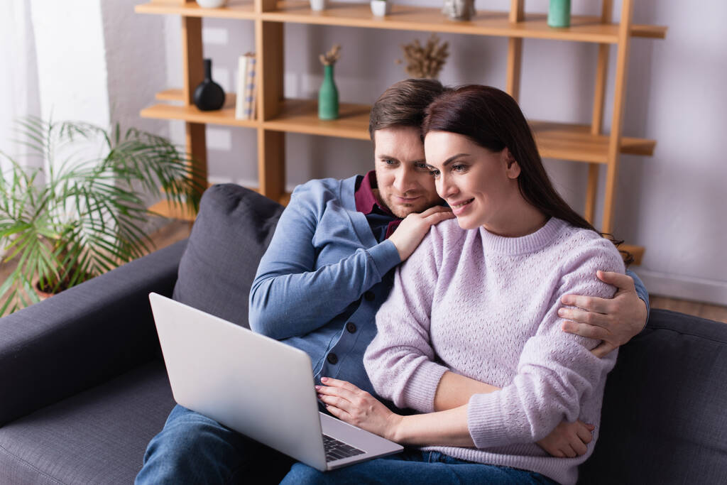 Улыбающаяся женщина смотрит на ноутбук рядом с мужем на диване  - Фото, изображение