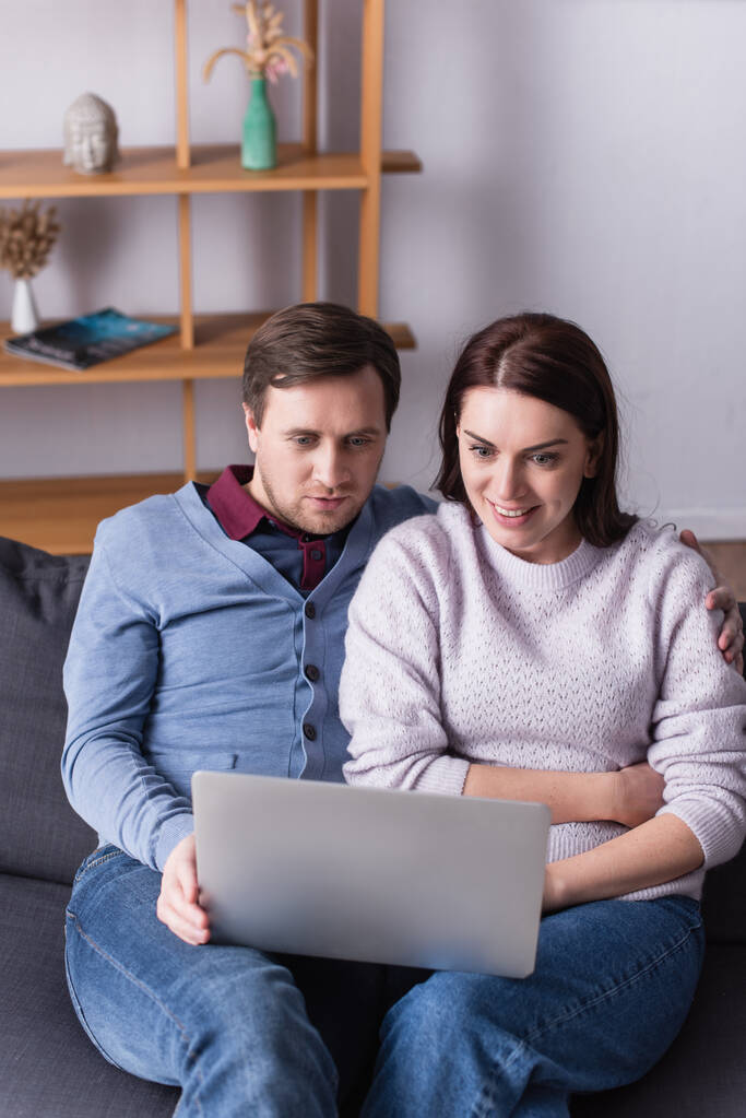 Hombre mirando el ordenador portátil cerca de esposa alegre en el sofá en la sala de estar  - Foto, Imagen