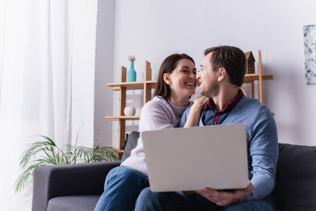 glimlachende vrouw kijken naar echtgenoot houden laptop op wazig voorgrond thuis  - Foto, afbeelding