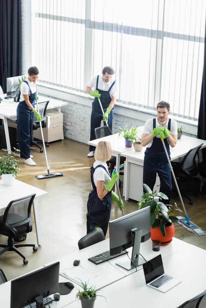 Ofisteki bilgisayarların yakınındaki yerleri temizleyen çok ırklı temizlikçilerin yüksek açılı manzarası  - Fotoğraf, Görsel