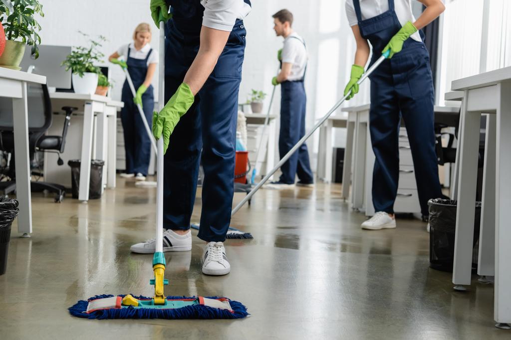 Limpiador de piso de limpieza con fregona cerca de colegas que trabajan sobre fondo borroso en la oficina  - Foto, Imagen