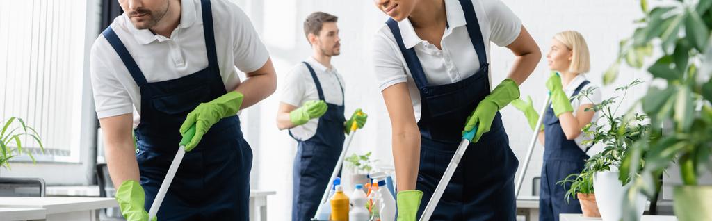 Limpiadores interraciales sosteniendo trapeadores cerca de colegas en un fondo borroso en la oficina, pancarta  - Foto, imagen