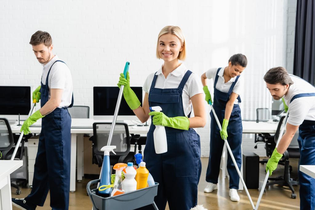 Усміхнений прибиральник тримає пральний порошок і мопед біля багатоетнічних колег в офісі
  - Фото, зображення