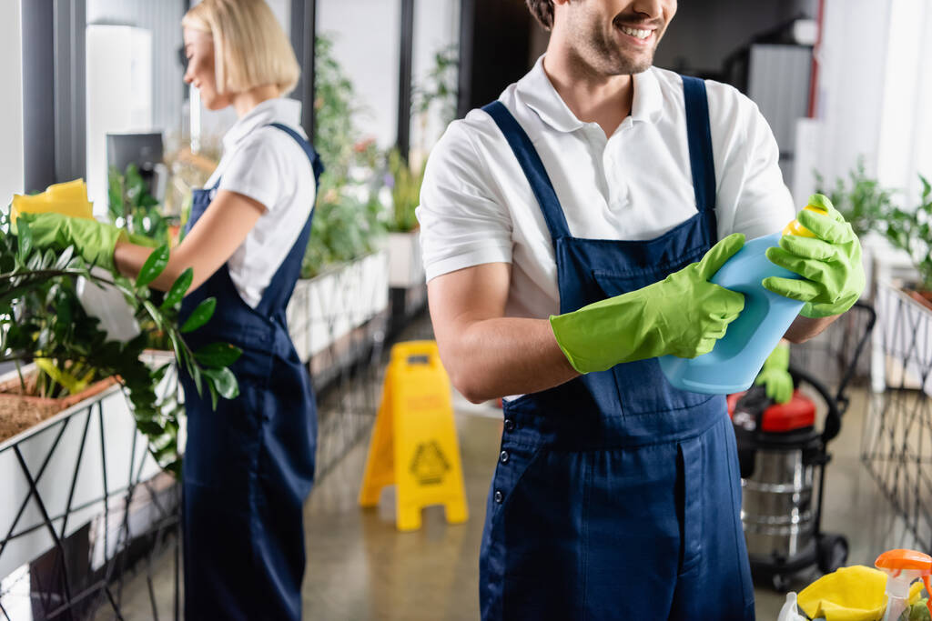 Улыбающийся чистильщик держит моющее средство рядом с коллегой на размытом фоне в офисе  - Фото, изображение