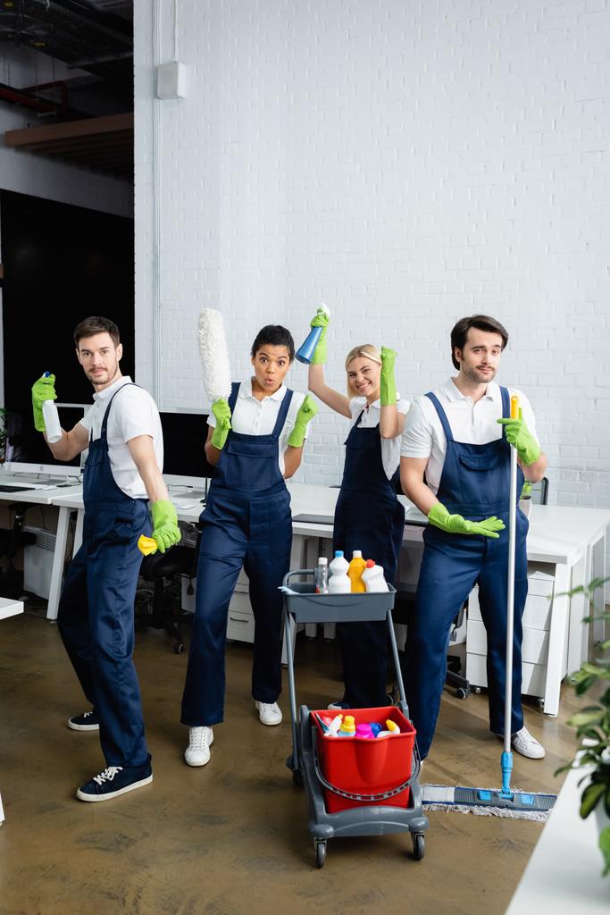 Limpiadores interraciales en overoles con artículos de limpieza en la oficina  - Foto, Imagen
