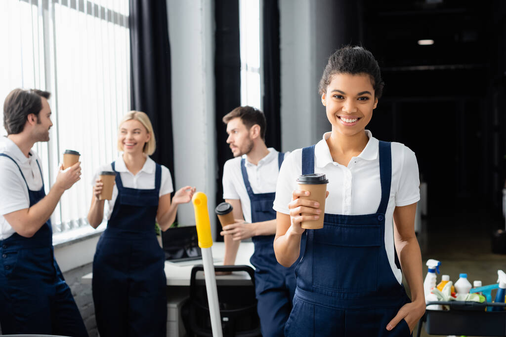 African american cleaner holding paper cup near colleagues on blurred background in office  - Photo, Image