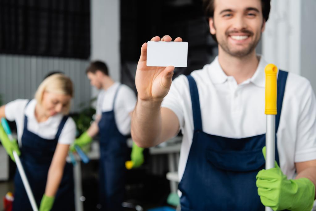 Empty card in hand of smiling cleaner with mop on blurred background in office  - Photo, Image